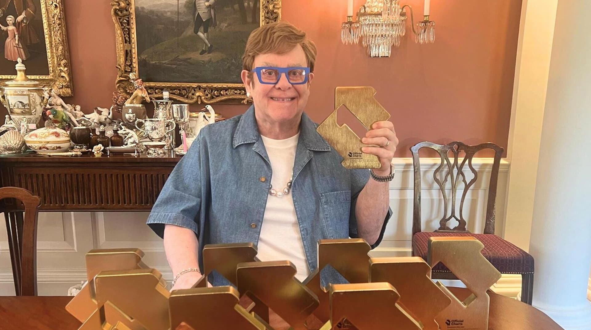 Elton John en chemise en jean, entouré de trophées dorés à table