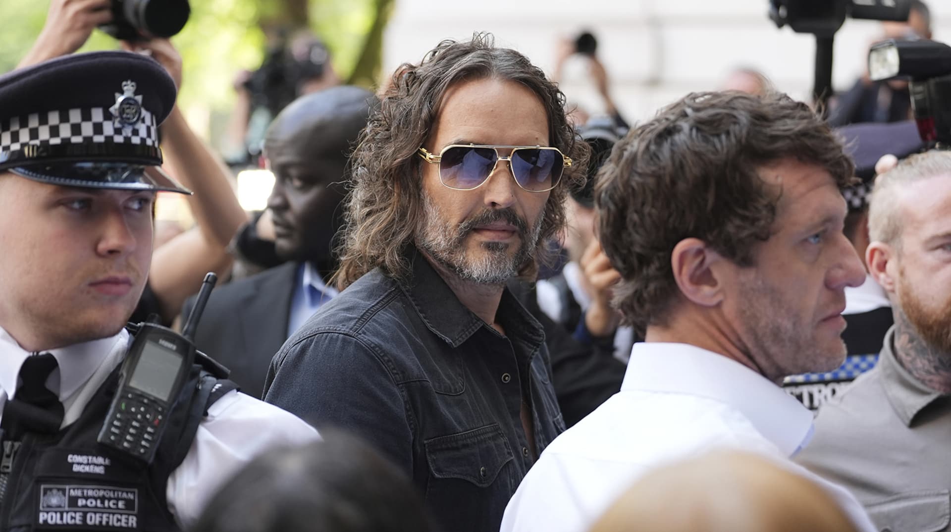 Russell Brand, lunettes de soleil, entouré de policiers et de photographes devant le tribunal