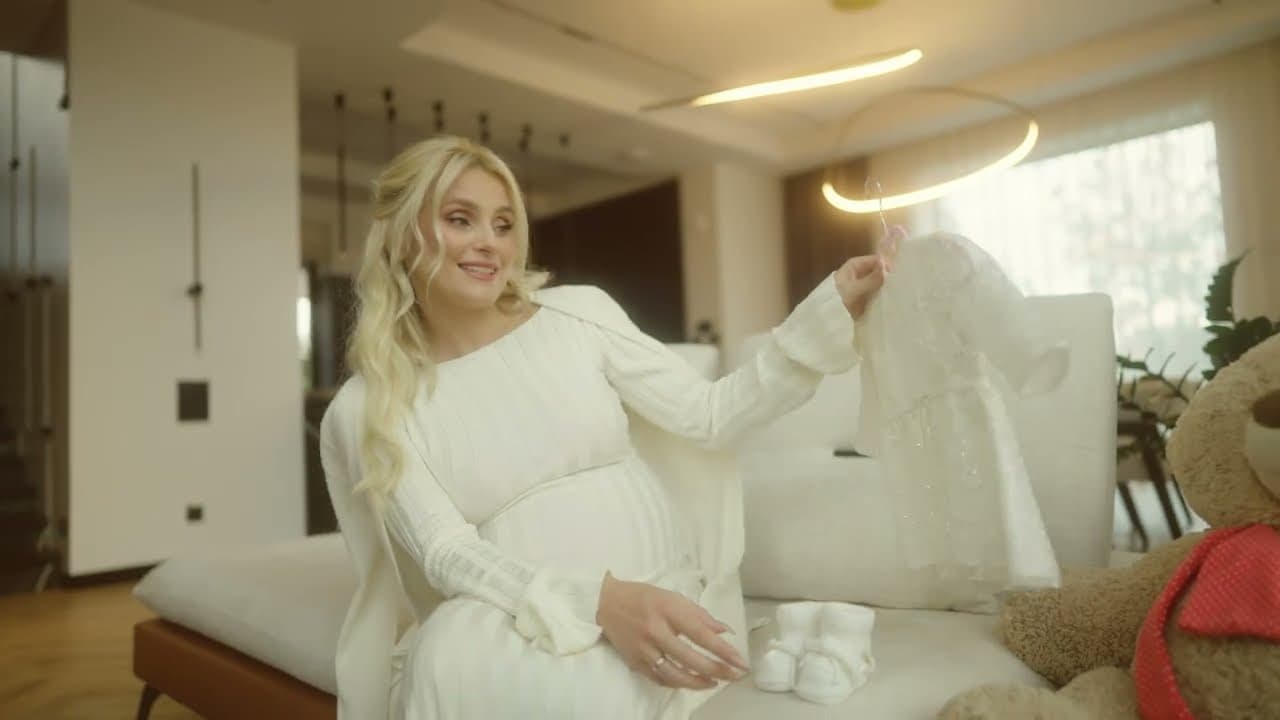 Iryna Fedyshyn en robe blanche, assise sur un canapé, regarde des vêtements de bébé à côté d’un ours en peluche