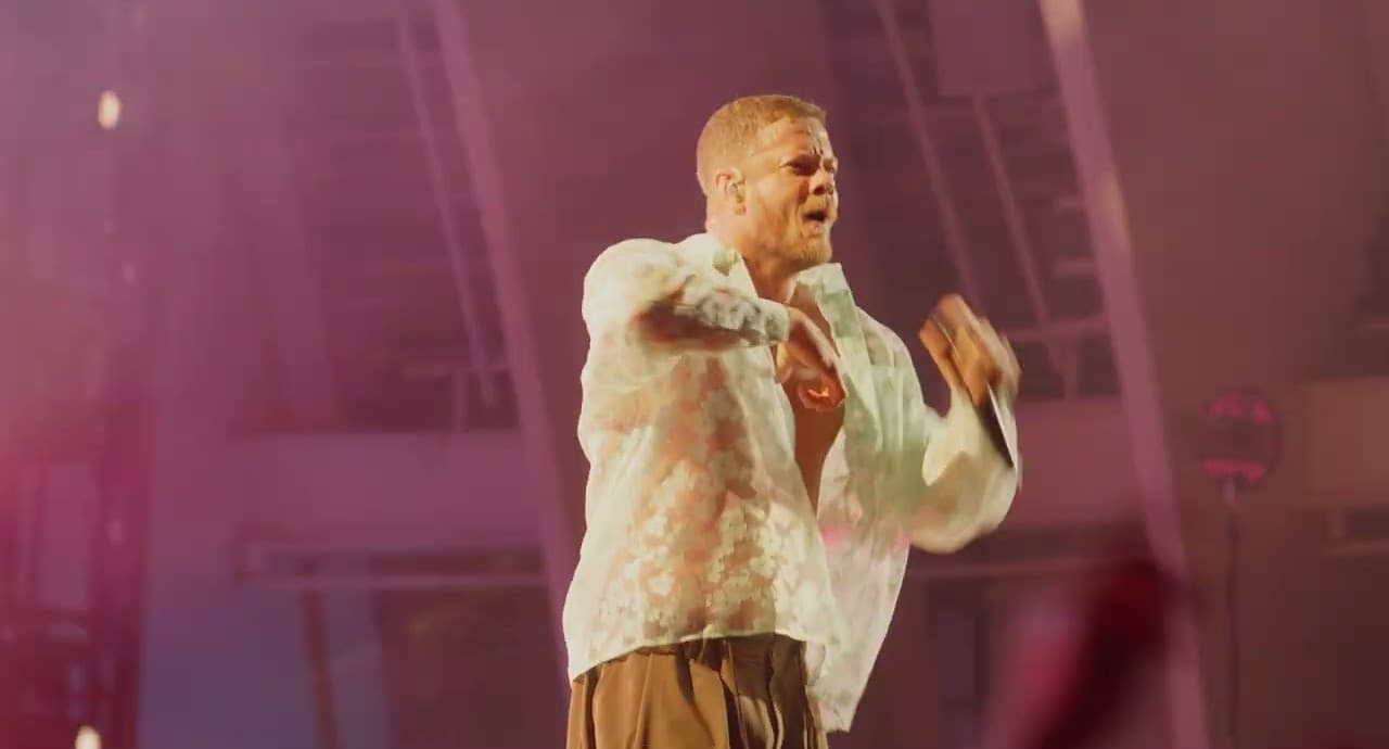 Dan Reynolds en chemise blanche en dentelle chante sur une scène baignée de lumière rose