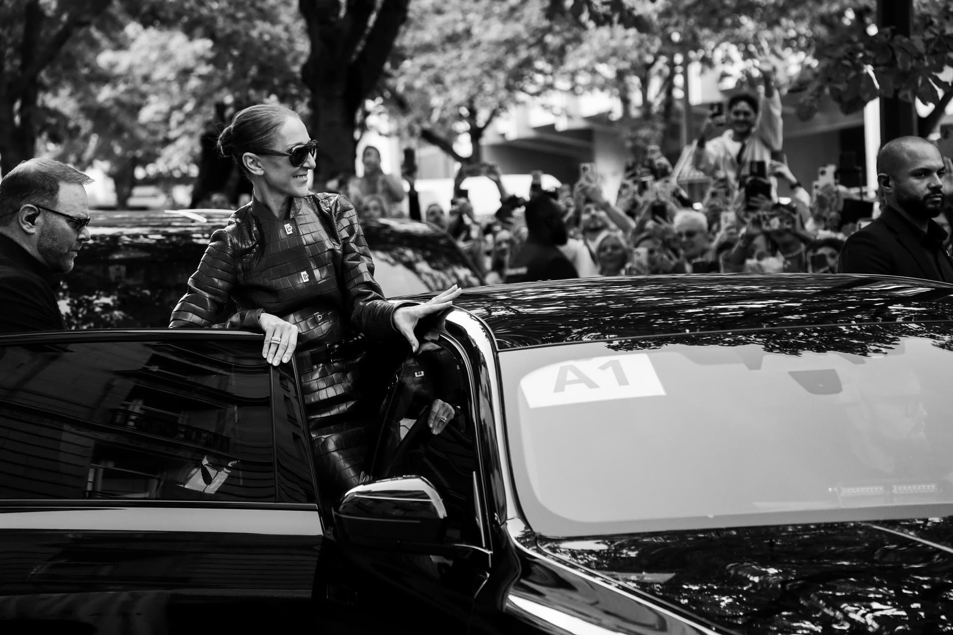 Photo en noir et blanc : Céline Dion en veste de cuir sort d’une voiture face à une foule d’admirateurs