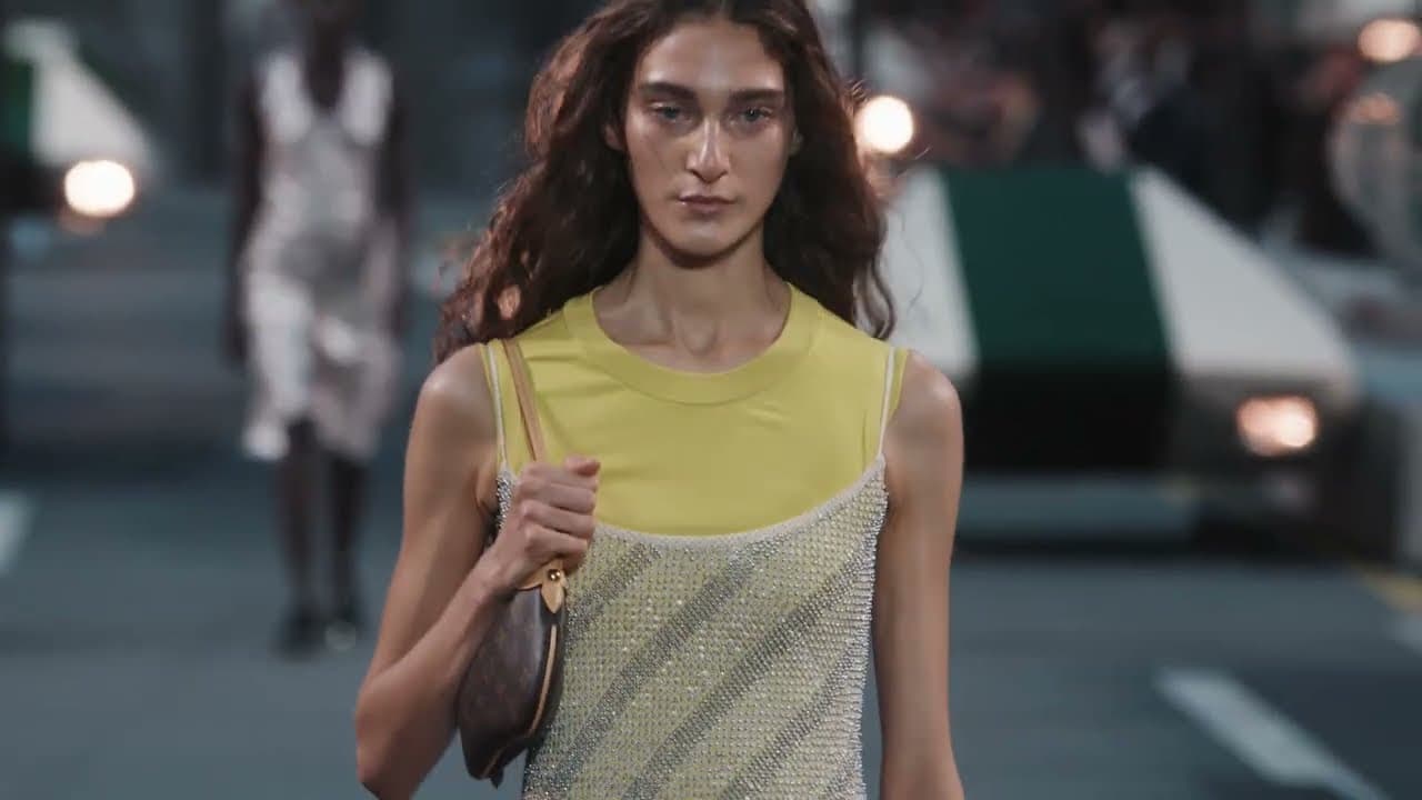 Модель у жовтому топі й сітчастій сукні з сумкою Louis Vuitton на подіумі в Луврі, позаду — летючі авто