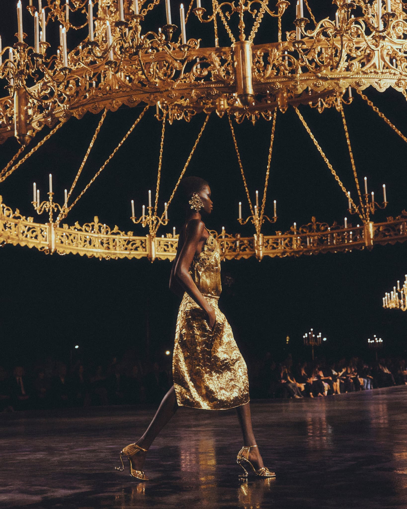 Mannequin Saint Laurent en robe dorée sur le podium, sous un immense lustre
