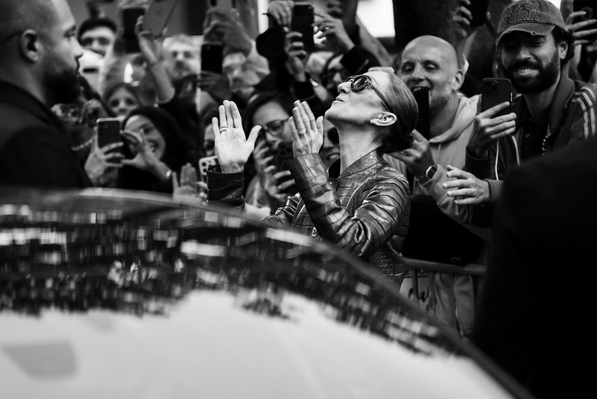 Photo en noir et blanc : Céline Dion, lunettes noires, sourit au milieu de fans munis de smartphones