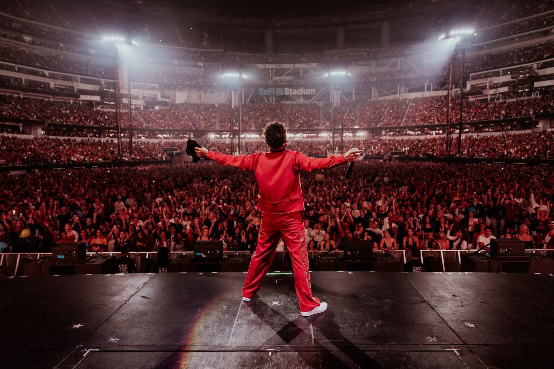 Bruno Mars en costume rouge, bras écartés sur scène devant un stade plein