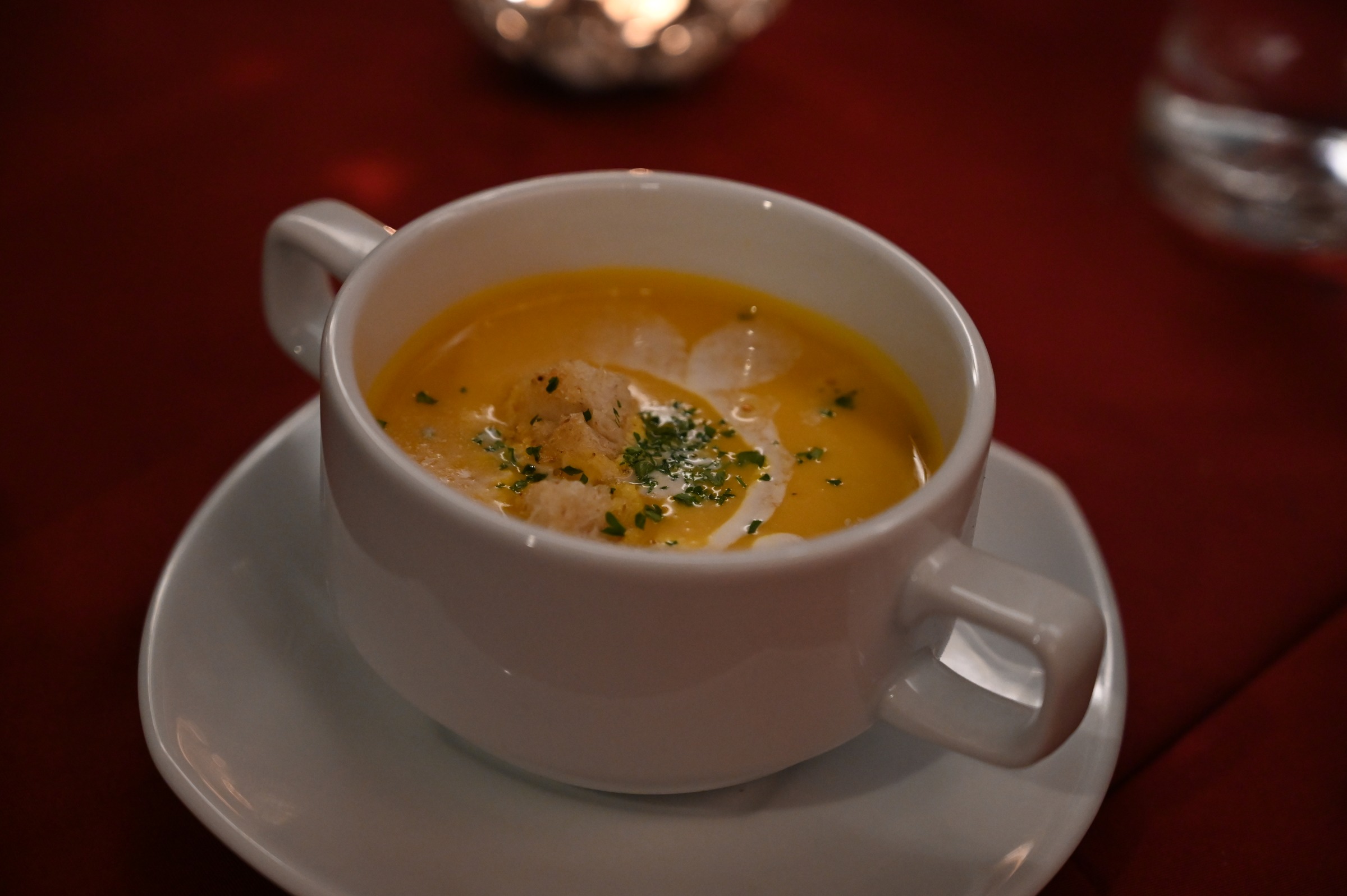 Velouté de courge avec une volute de crème et des herbes, dans une tasse blanche sur une nappe rouge