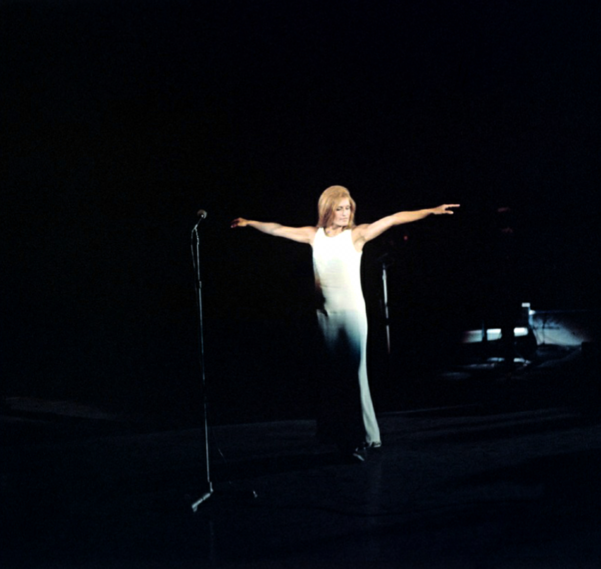 Dalida en longue robe claire, bras écartés, sur une scène sombre près d’un micro