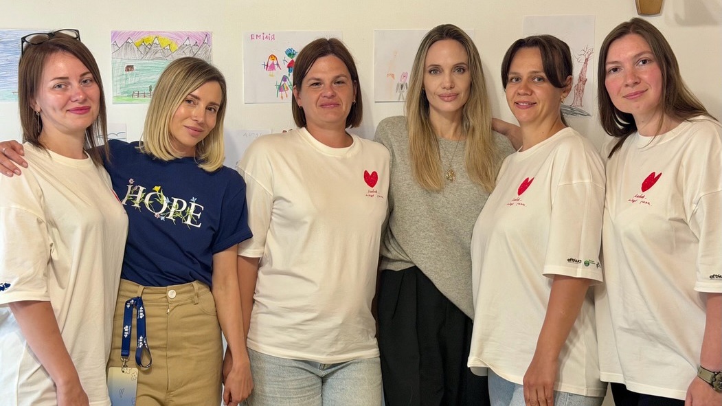 Анджеліна Джолі стоїть у центрі між пʼятьма жінками у білих футболках із червоним серцем на тлі дитячих малюнків