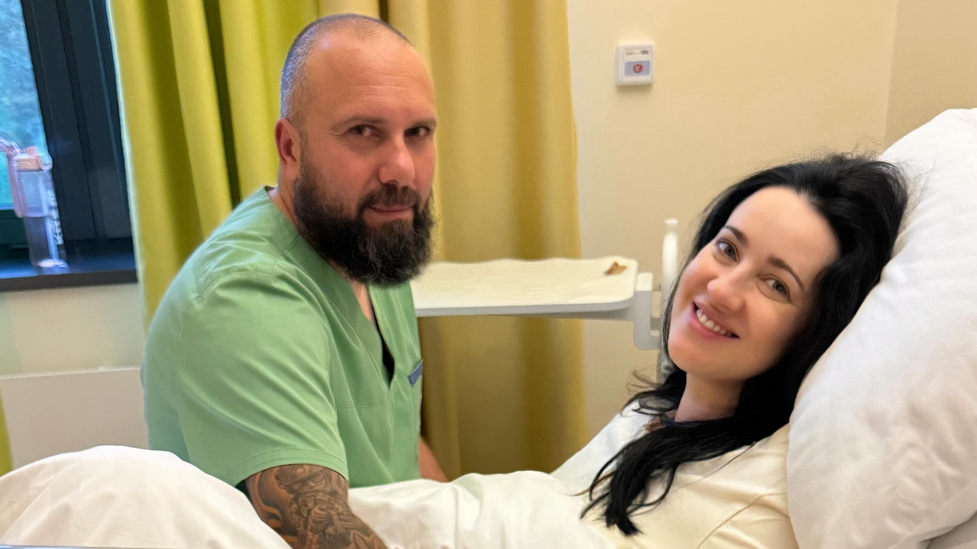 Соломія Вітвіцька в ліжку в пологовій палаті, поруч сидить її чоловік Олексій Ситайло в зеленому медичному костюмі