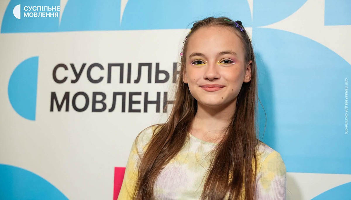 Усміхнена Еріка Колесніченко з довгим русявим волоссям на тлі логотипа Суспільного Мовлення