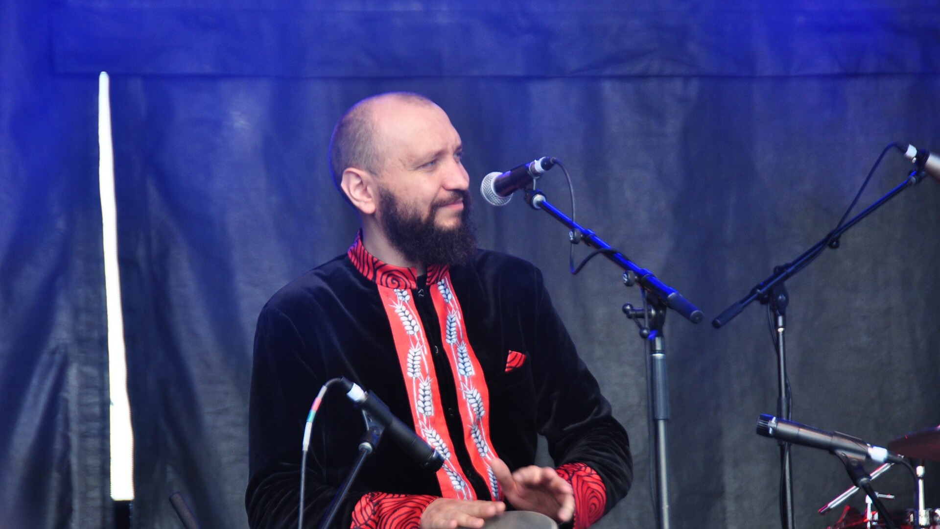 Марко Галаневич із DakhaBrakha в чорній оксамитовій сорочці з червоною вишивкою грає на барабані біля мікрофона на тлі синьої завіси