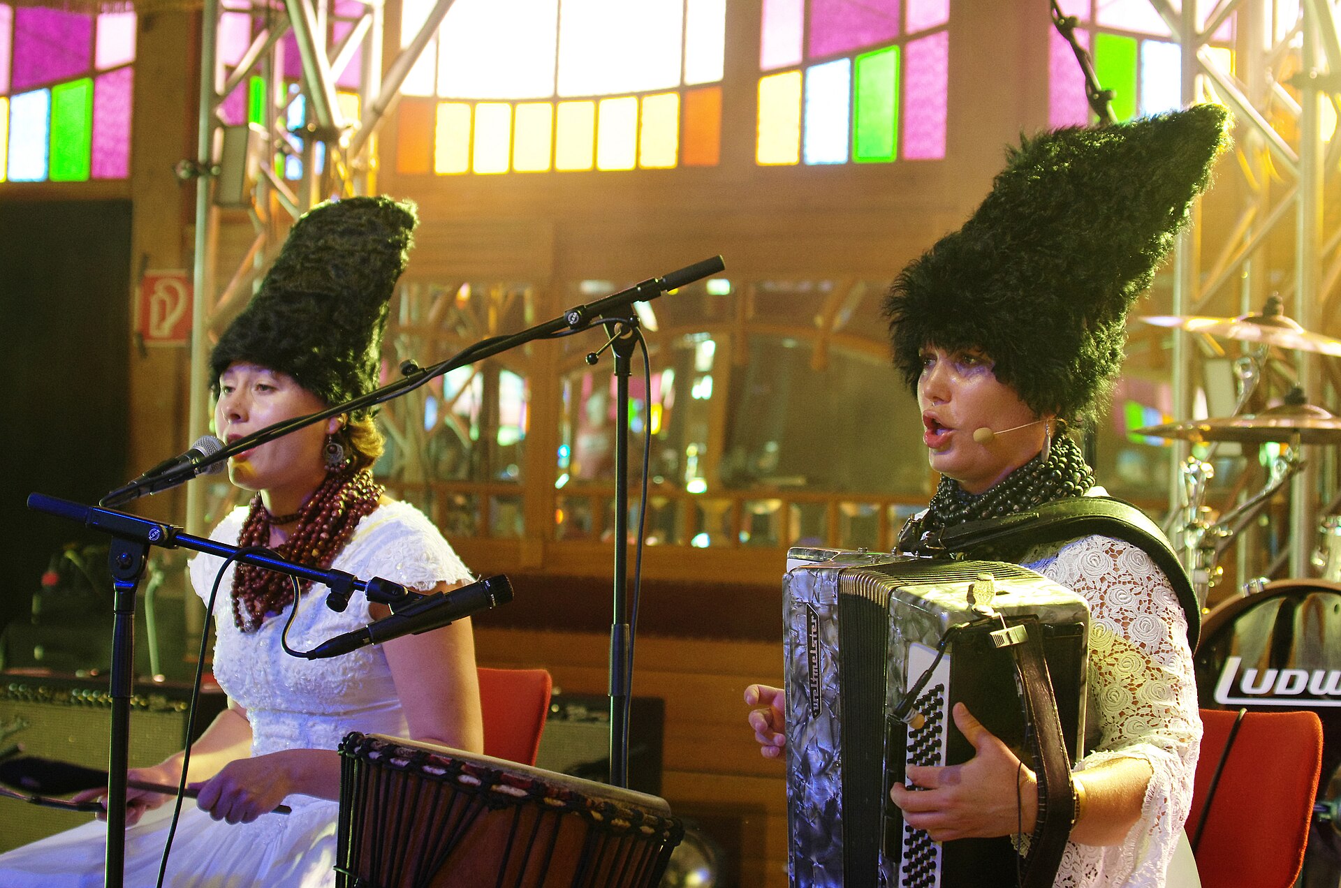 Дві учасниці DakhaBrakha у високих чорних шапках співають на сцені, одна грає на акордеоні, позаду кольорові вітражні вікна