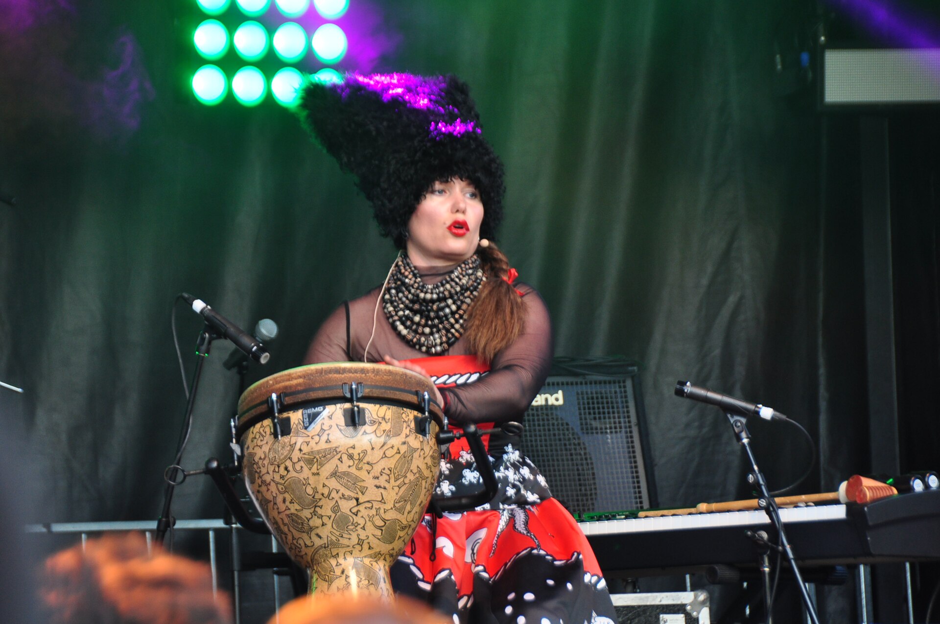 Ірина Коваленко з DakhaBrakha у високій шапці й намисті грає на джембе на сцені із зеленим світлом